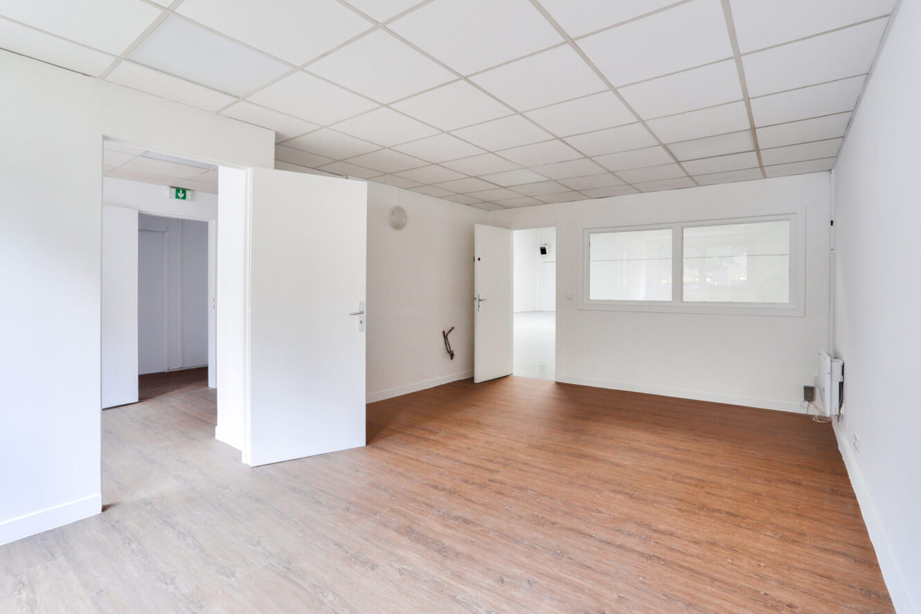 Bureau vide avec des murs blancs, des portes ouvertes, une fenêtre intérieure, un sol en bois et un plafond carrelé avec des lumières.