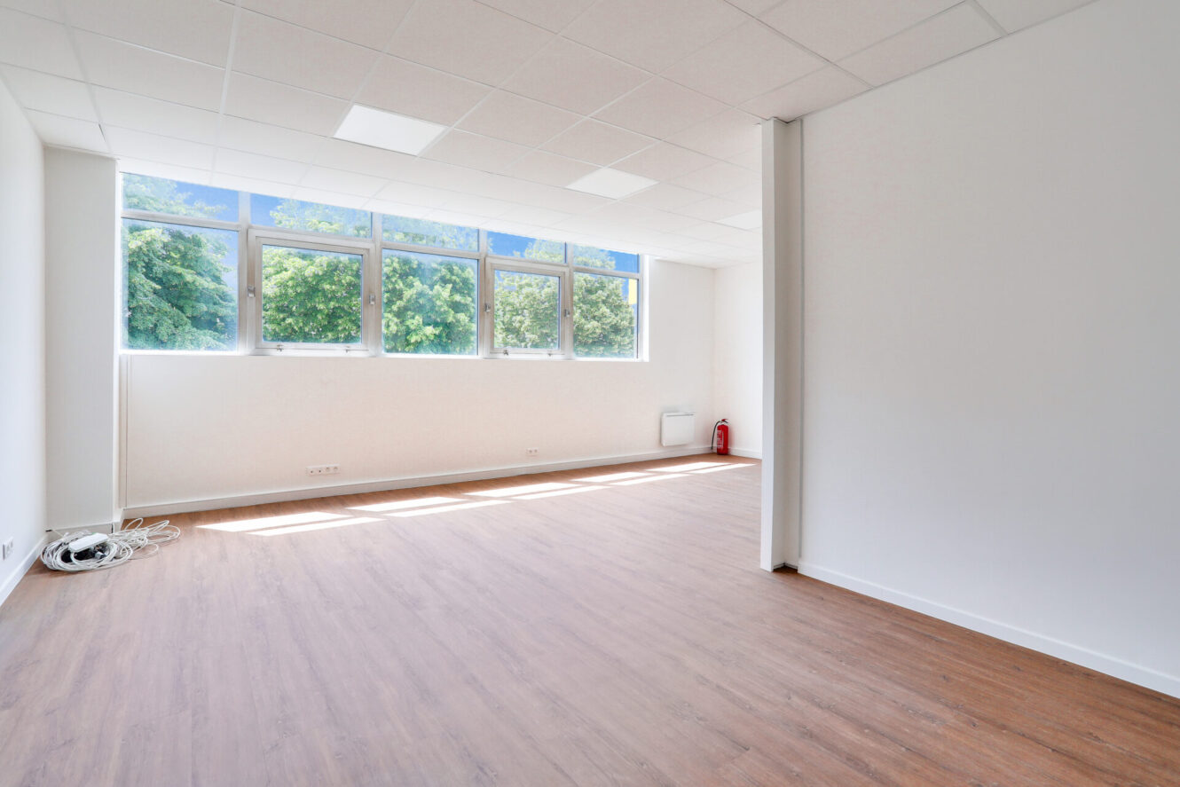 Bureau vide avec de grandes fenêtres, des murs blancs, des planchers en bois, des plafonniers, un extincteur et des câbles enroulés près du mur.
