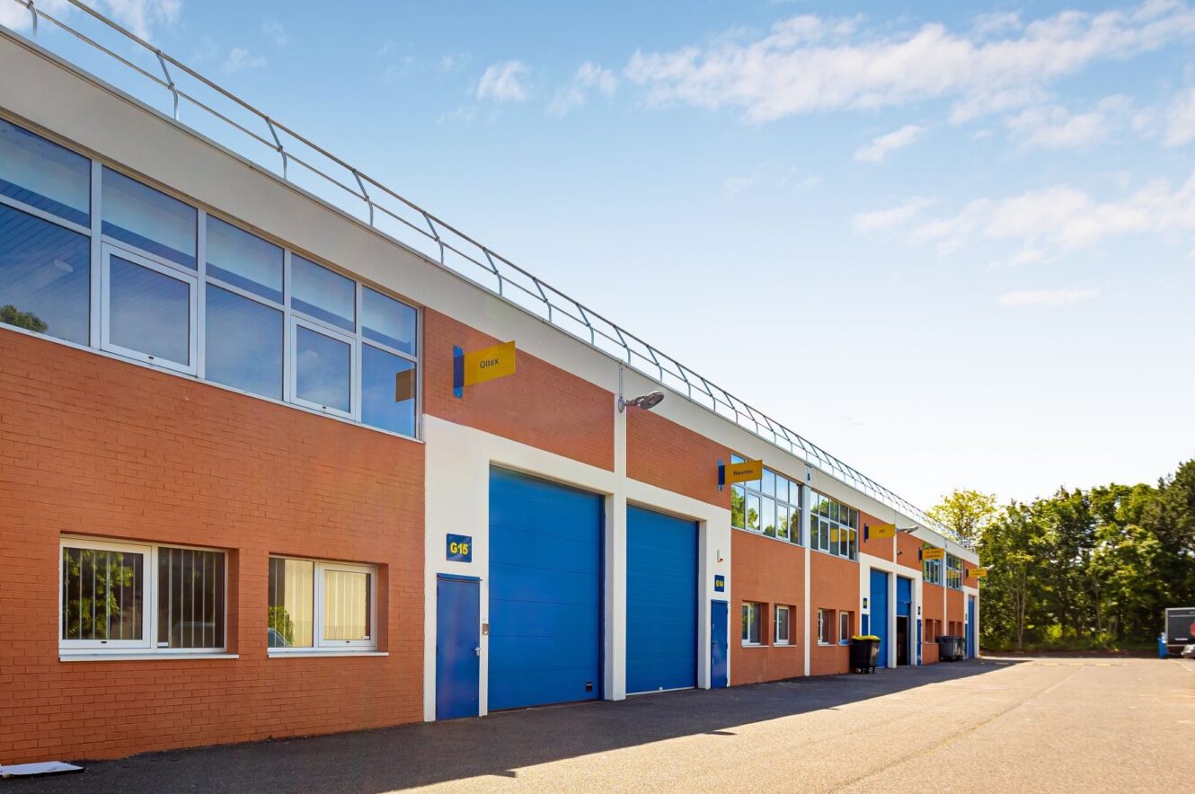 Une rangée d'entrepôts industriels aux portes bleues et aux enseignes jaunes, dotés de grandes fenêtres, sous un ciel dégagé.