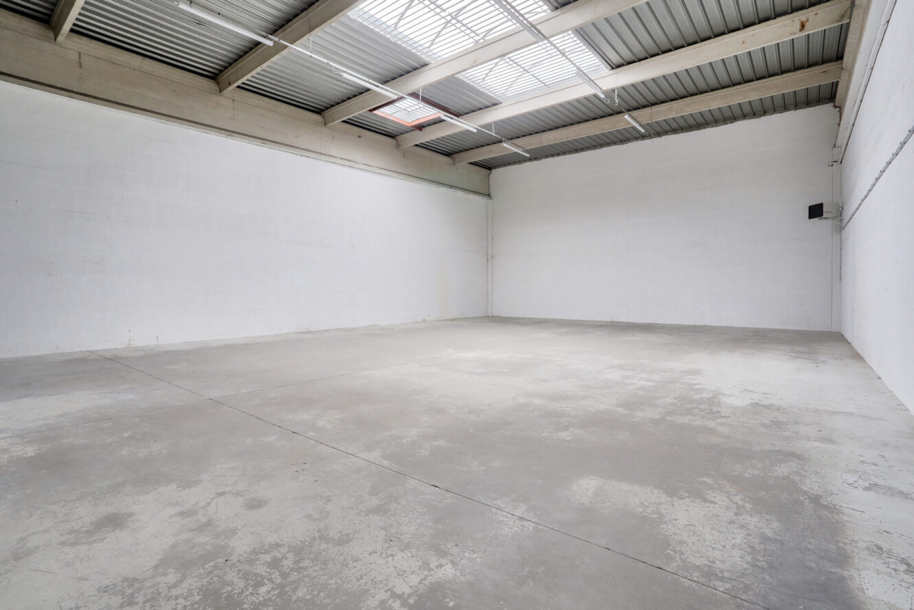 Entrepôt industriel vide avec des murs blancs, un sol en béton et des lucarnes dans un toit métallique qui laissent entrer la lumière naturelle dans l'espace ouvert.