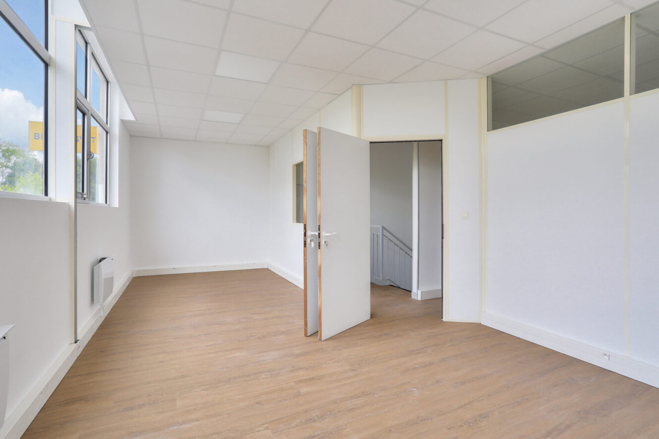 Pièce de bureau vide avec des murs blancs, des planchers en bois, de grandes fenêtres et une porte ouverte donnant sur un couloir avec des escaliers ; pas de meubles ni de personnes.