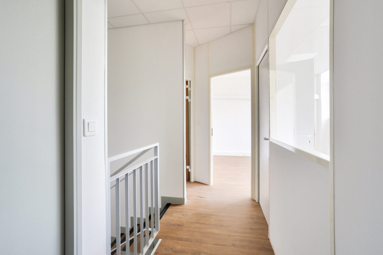 Couloir lumineux avec des murs blancs, un sol en bois, une rampe d'escalier et des portes ouvertes sur des pièces baignées de lumière naturelle.