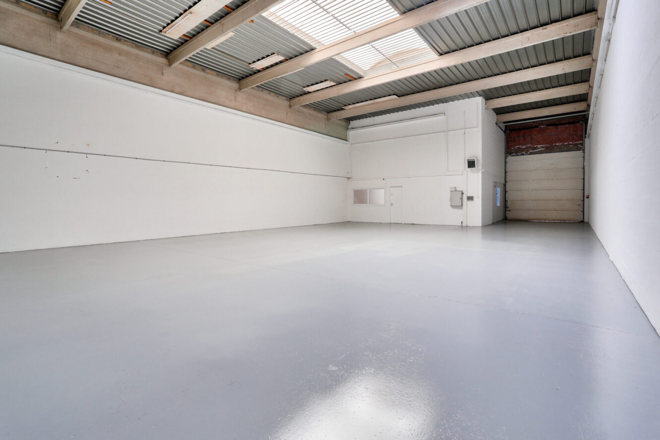 Entrepôt industriel vide avec des murs blancs, un haut plafond, des puits de lumière, un sol en béton, une grande porte à volets et des installations minimales.