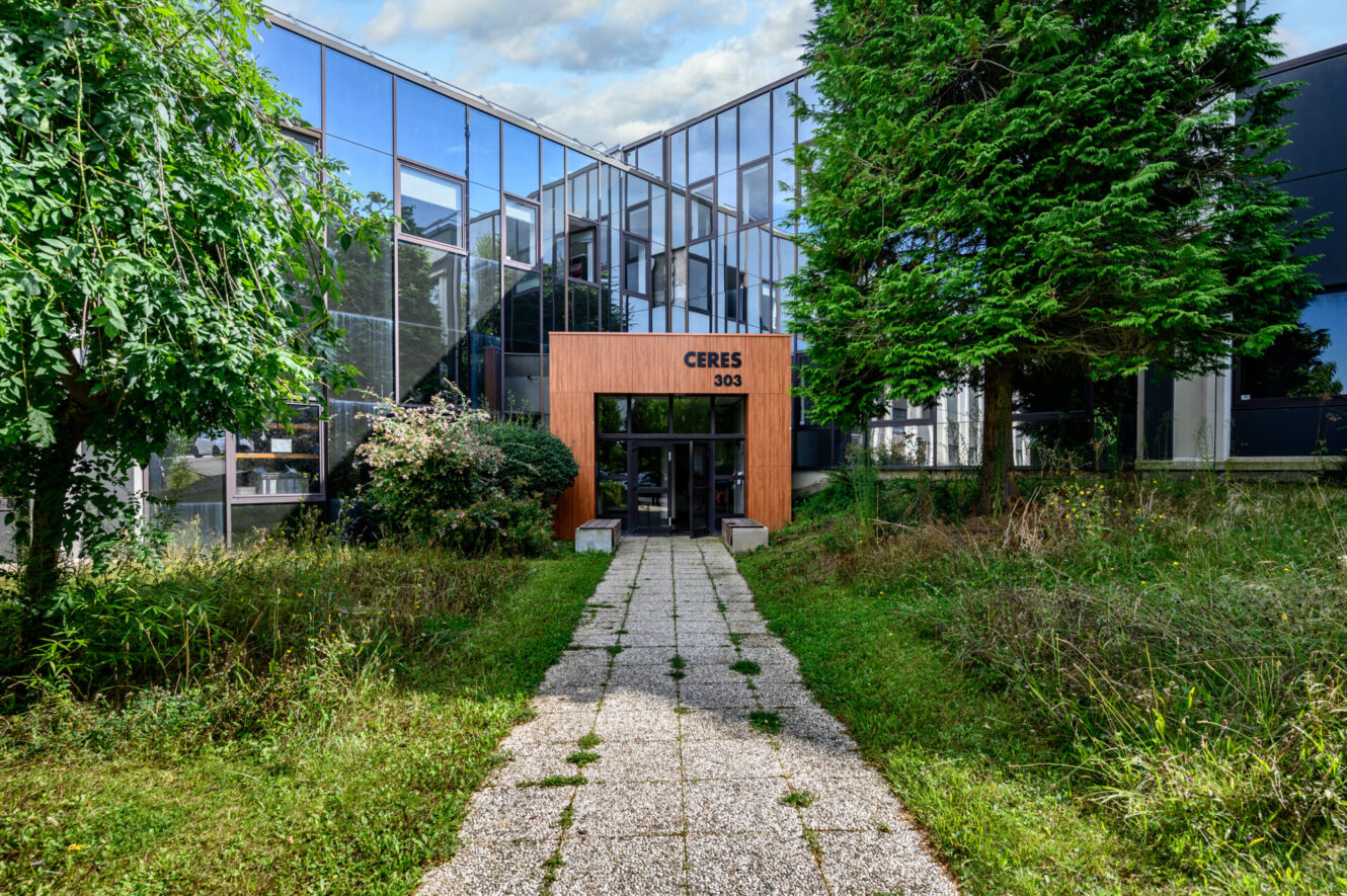 Un immeuble de bureaux en verre avec une entrée en bois étiquetée CERES 303, de l'herbe et des arbres envahissants tout autour, et une allée menant à la porte.