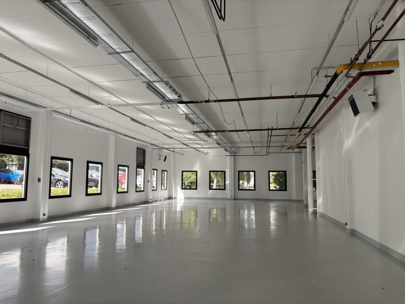 Pièce vide de style industriel avec des sols brillants, des murs blancs, plusieurs fenêtres et des tuyaux de plafond apparents. La lumière du soleil entre à flots.
