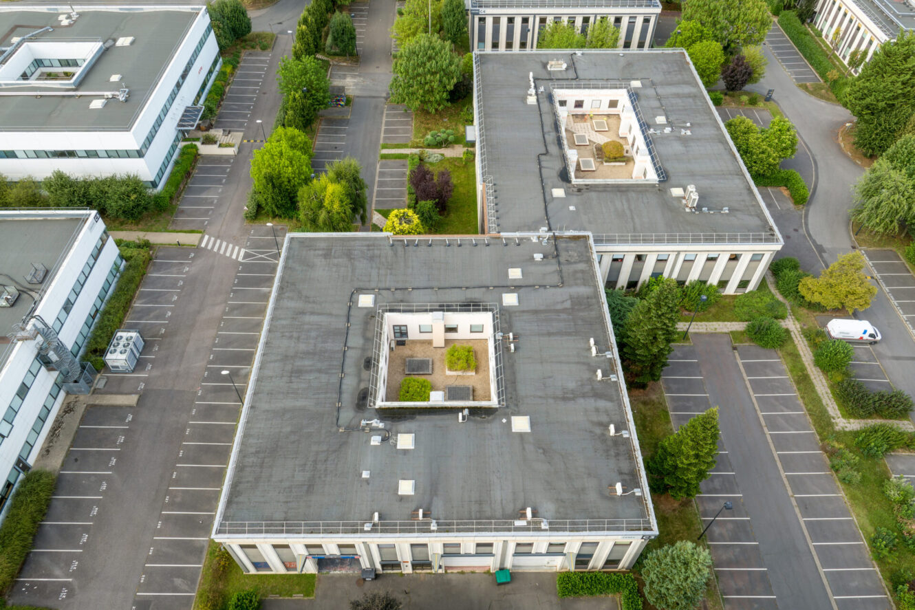 Vue aérienne de trois immeubles de bureaux aux toits plats, entourés de verdure et de parkings en grande partie vides.