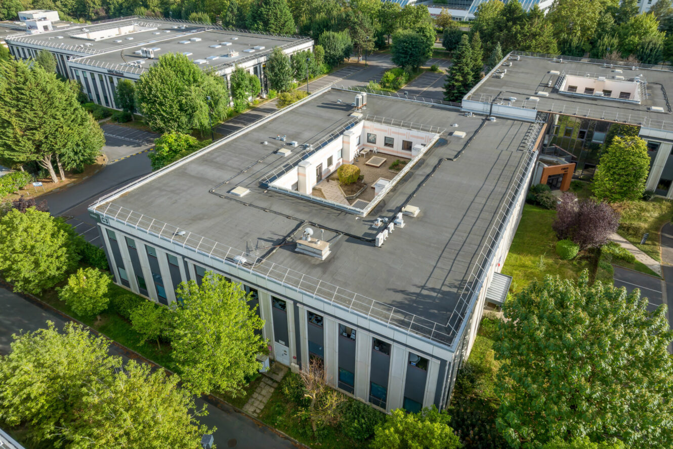 Vue aérienne d'un immeuble de bureaux moderne rectangulaire à toit plat, entouré d'arbres, de bâtiments voisins et de parkings.