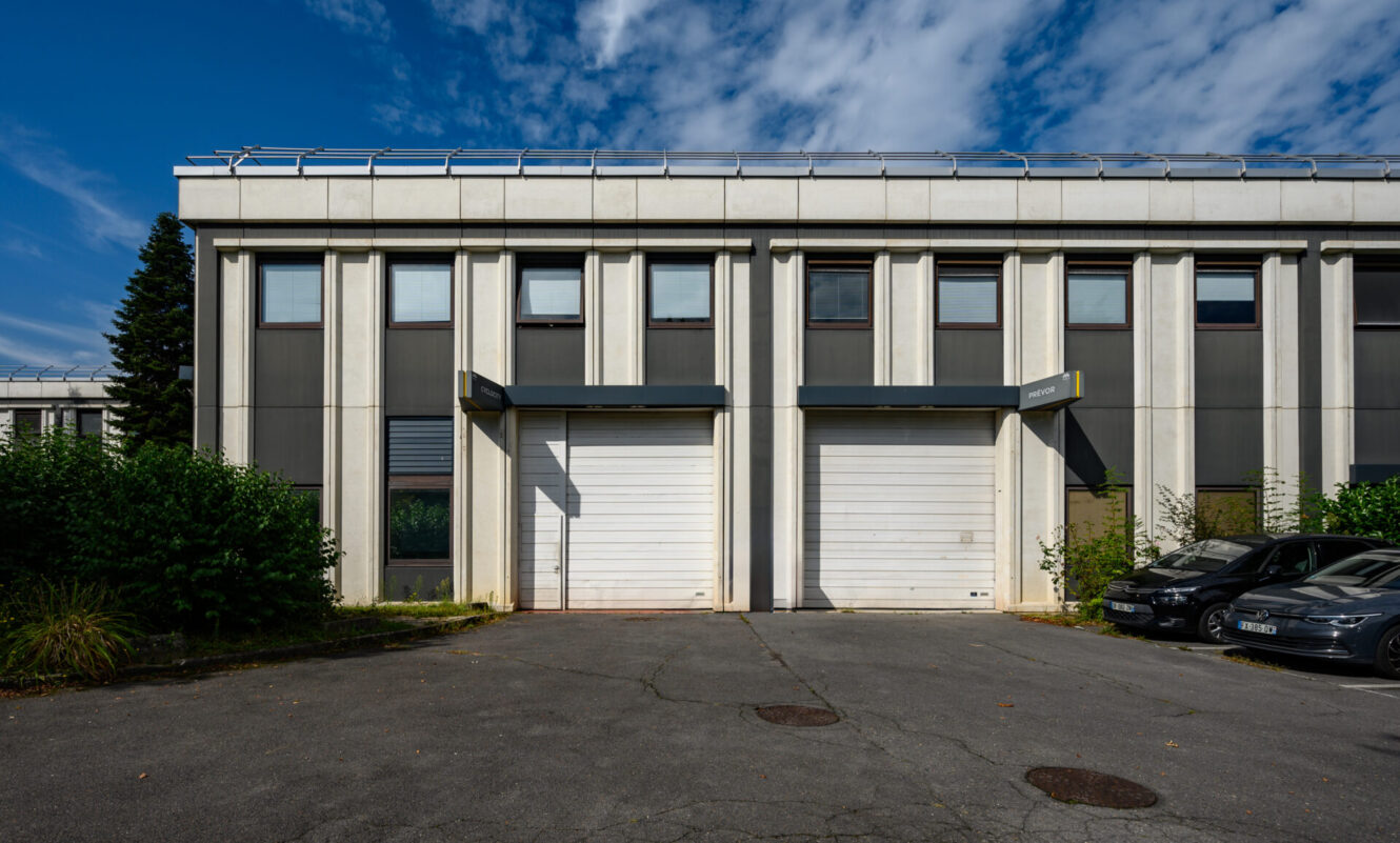 Extérieur d'un bâtiment industriel moderne avec deux portes de garage blanches fermées, de hautes fenêtres au-dessus, deux voitures garées sur la droite et un ciel partiellement nuageux.