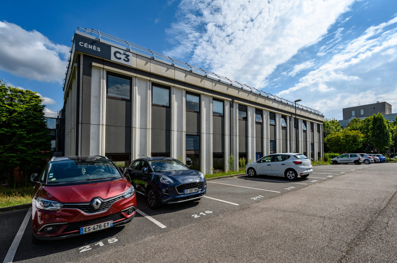 Par une journée ensoleillée et partiellement nuageuse, trois voitures sont garées sur des places numérotées à l'extérieur d'un immeuble de bureaux moderne portant le nom de CÉRÈS C3.