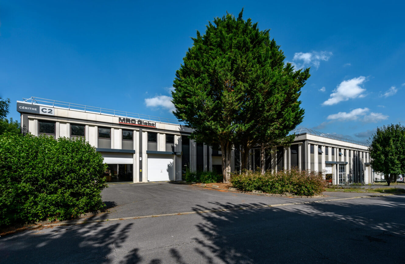 Un immeuble de bureaux moderne de deux étages avec des colonnes blanches et de grandes fenêtres, partiellement caché par des arbres et des buissons sous un ciel bleu clair.