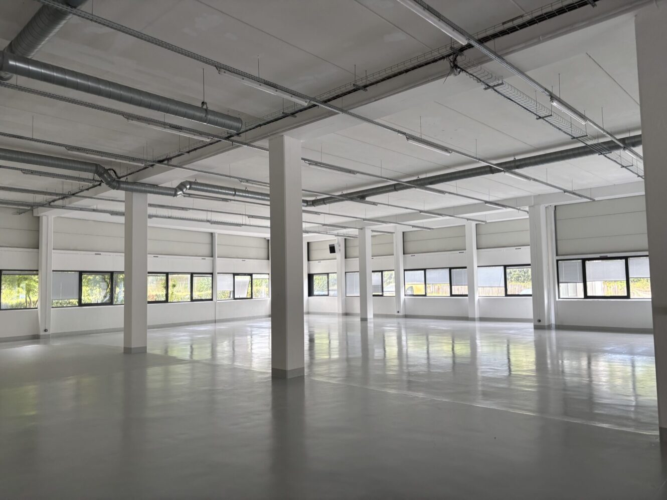 Intérieur industriel ou commercial spacieux et vide, avec des murs blancs, de grandes fenêtres, des sols polis et des conduits apparents au plafond.