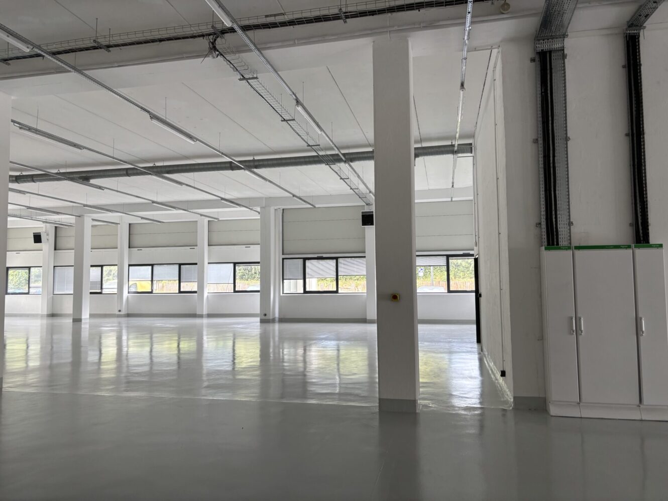 Grand entrepôt industriel vide aux murs blancs, à l'éclairage zénithal, aux conduits exposés et aux fenêtres sur un côté.