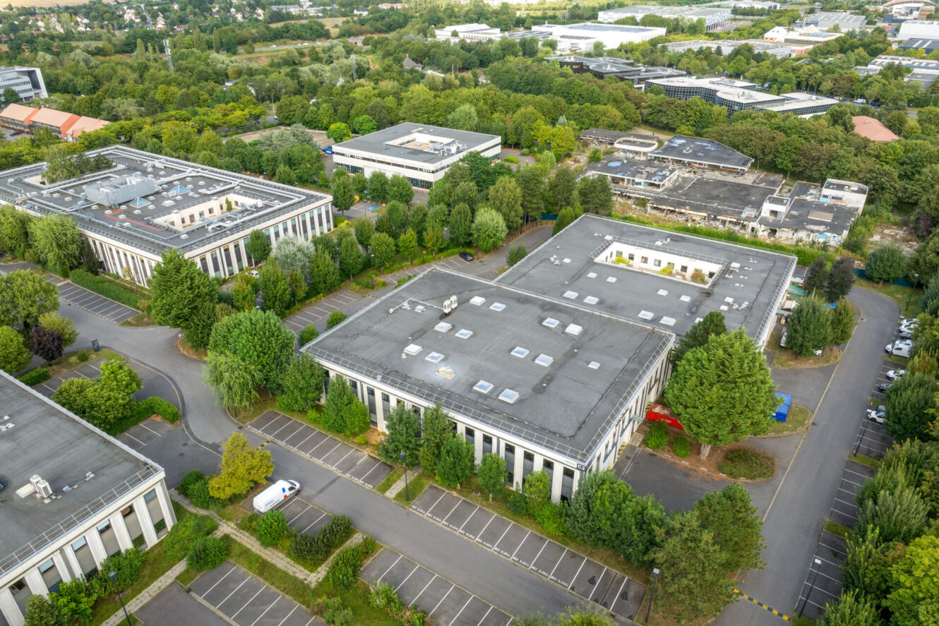 Vue aérienne d'immeubles de bureaux aux toits plats, entourés de parkings, d'arbres et d'espaces verts dans un parc d'activités.