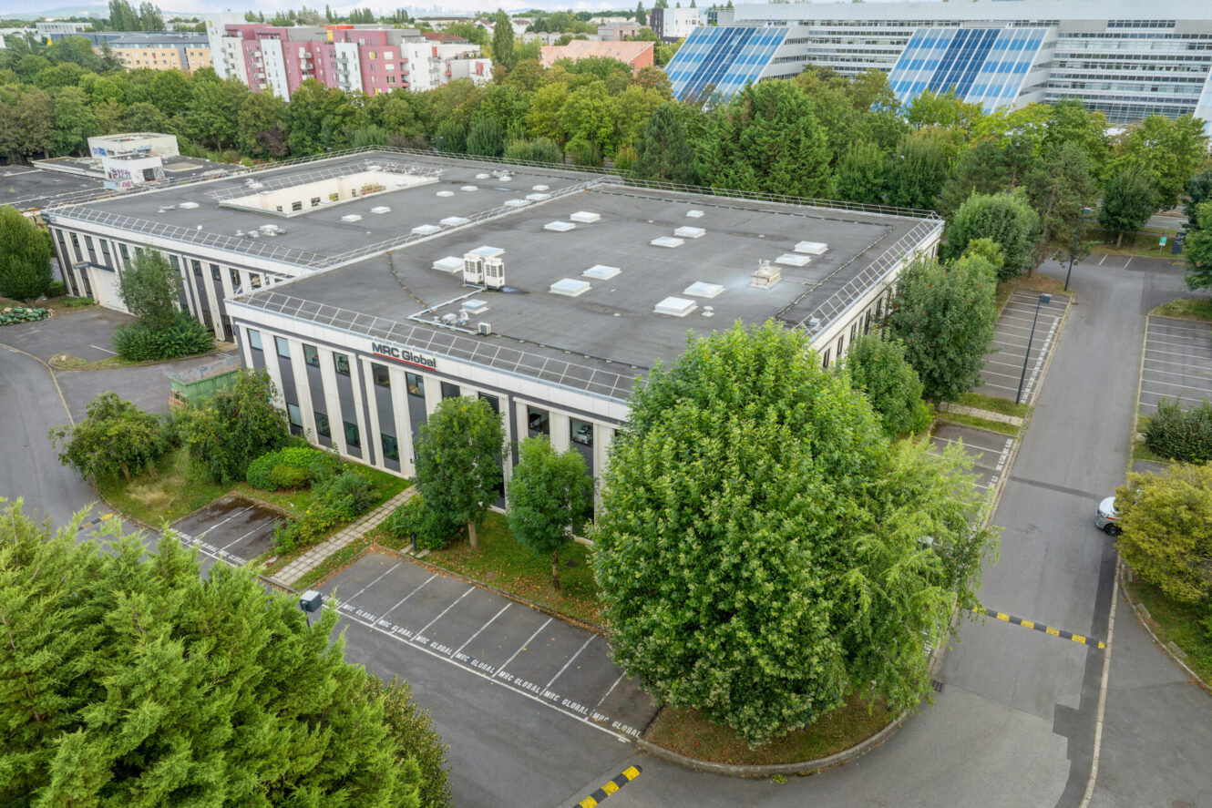 Vue aérienne d'un grand immeuble de bureaux rectangulaire avec les arbres environnants, les parkings et les habitations et entreprises voisines.