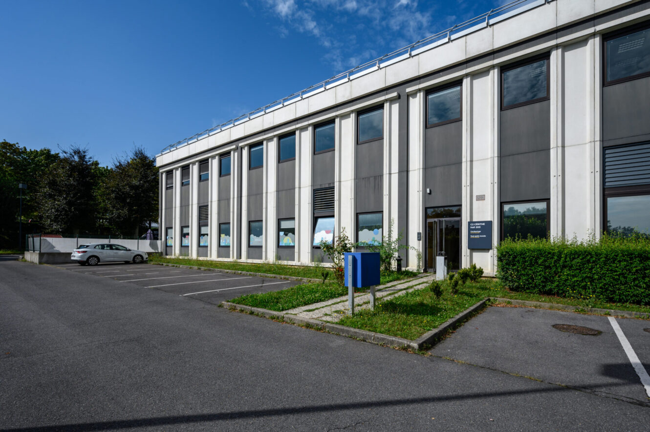 Un immeuble de bureaux de deux étages à l'extérieur gris et blanc, avec de grandes fenêtres, un parking à proximité et un téléphone d'urgence bleu à l'avant.