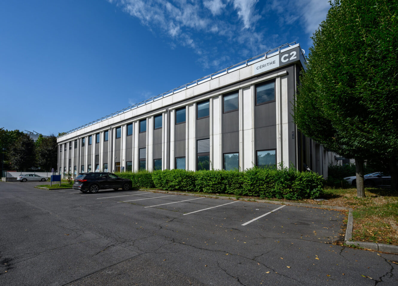 Immeuble de bureaux de deux étages étiqueté C2 Centre avec de hautes fenêtres, de la verdure et un parking presque vide sous un ciel bleu.