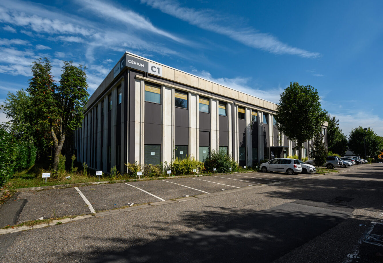 Un immeuble de bureaux moderne de deux étages étiqueté C1 avec des voitures garées et un parking vide, entouré d'arbres sous un ciel bleu.