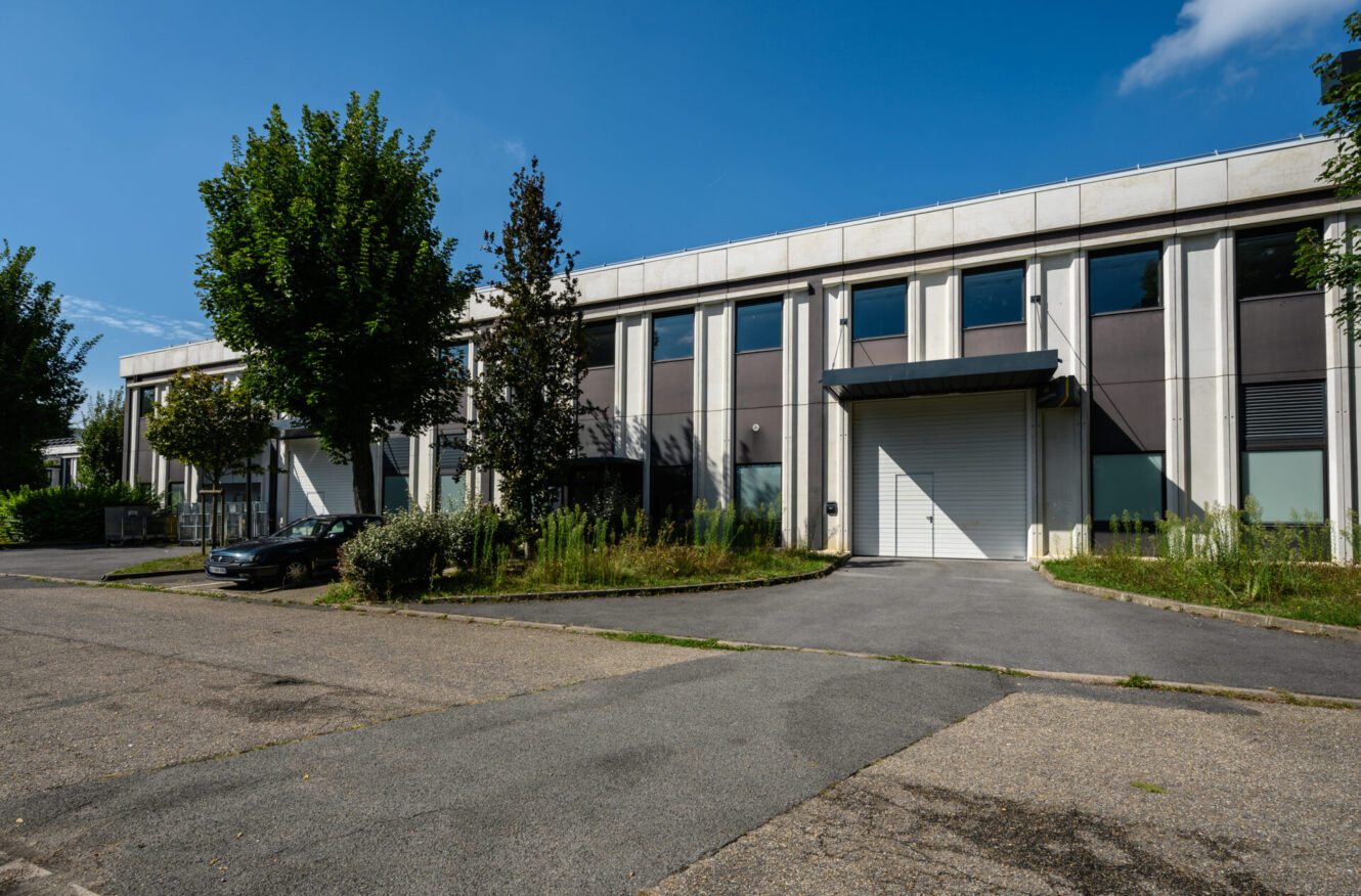 Bâtiment industriel ou de bureaux moderne à deux étages avec de grandes fenêtres, une porte de garage blanche à enroulement, des arbres et une voiture garée à l'extérieur sous un ciel bleu.