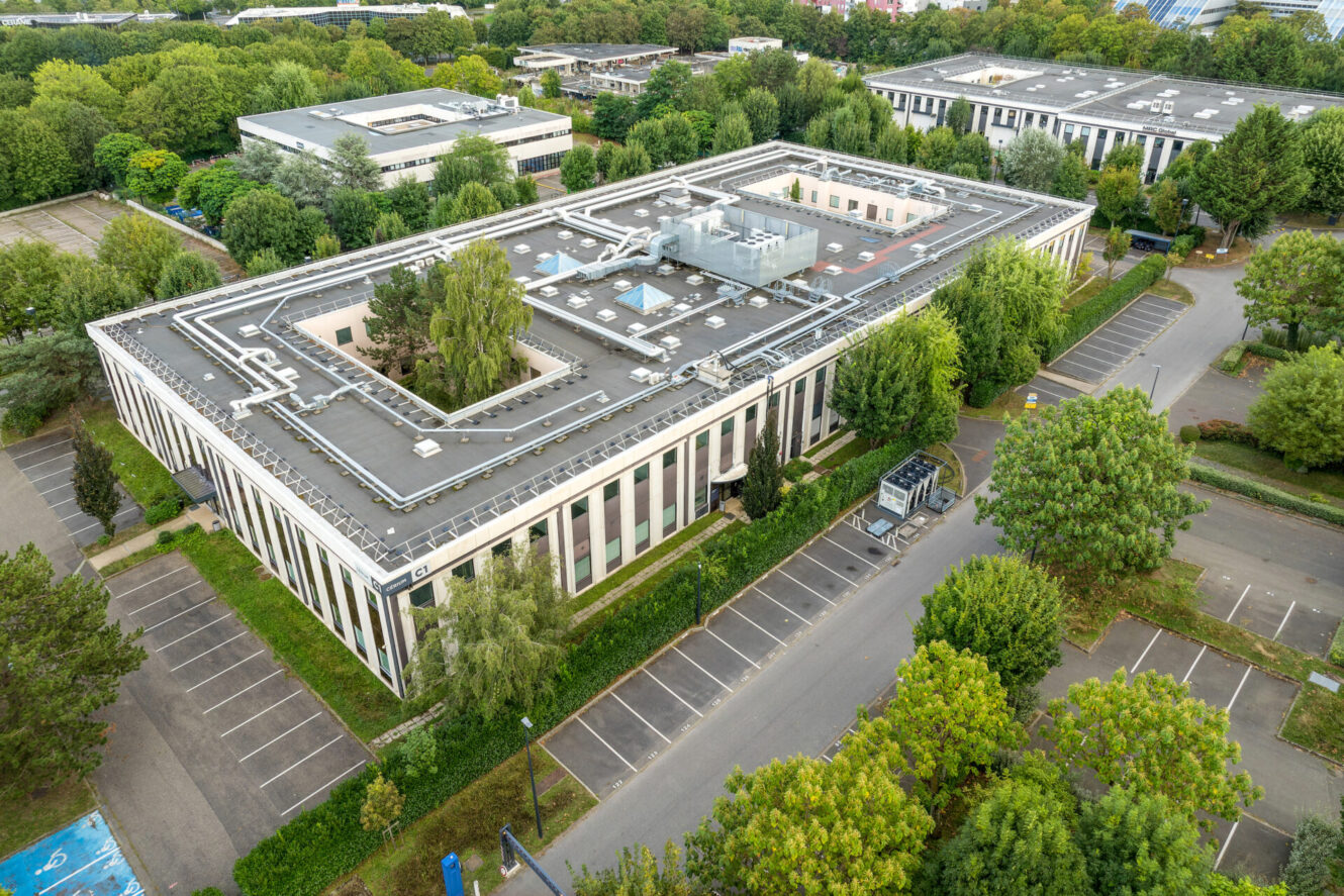 Vue aérienne d'un grand immeuble de bureaux rectangulaire à toit plat, entouré d'arbres et de parkings en grande partie vides.