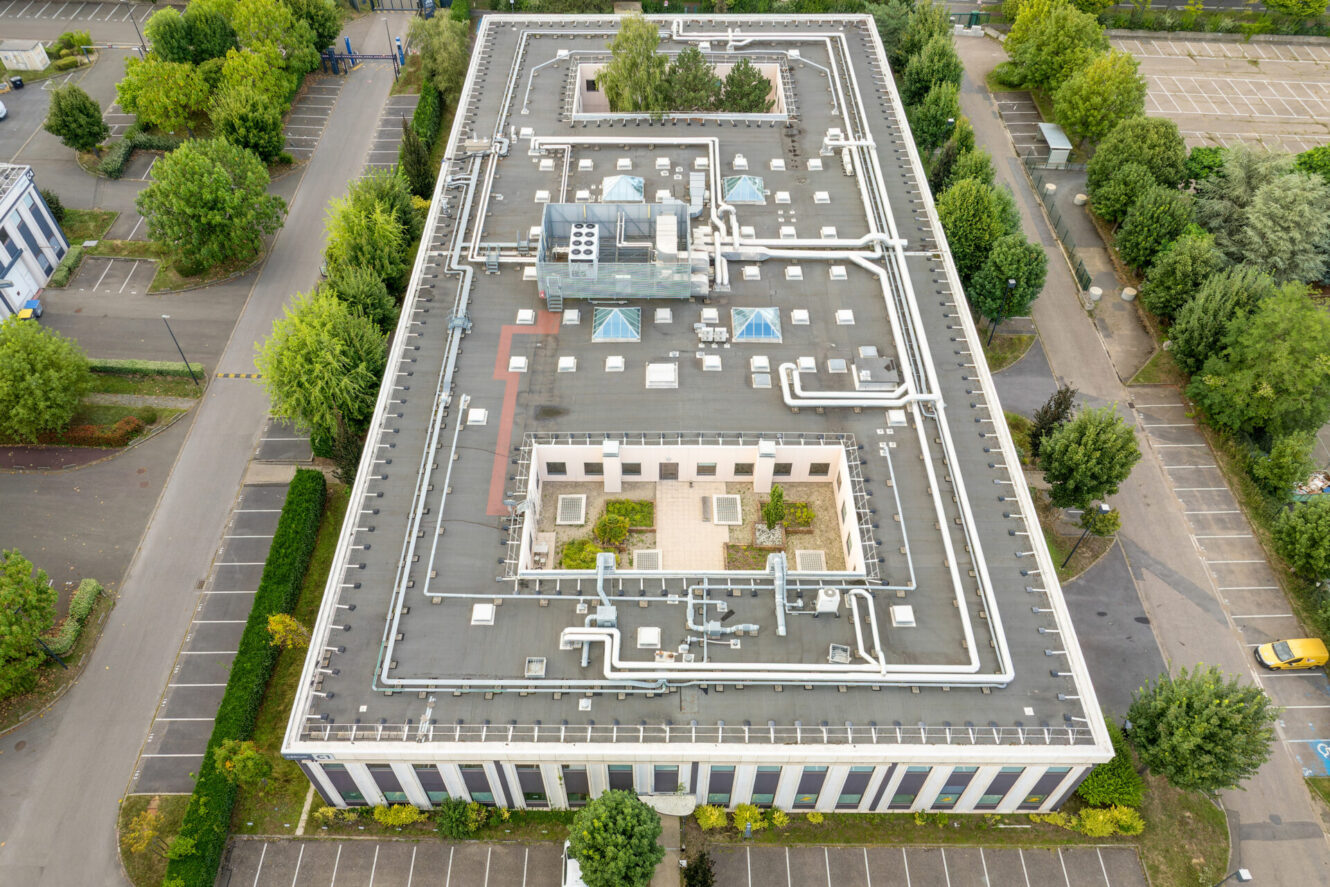 Vue aérienne d'un bâtiment rectangulaire à toit plat avec des tuyaux sur le toit, entouré d'arbres et de parkings avec des voitures garées.