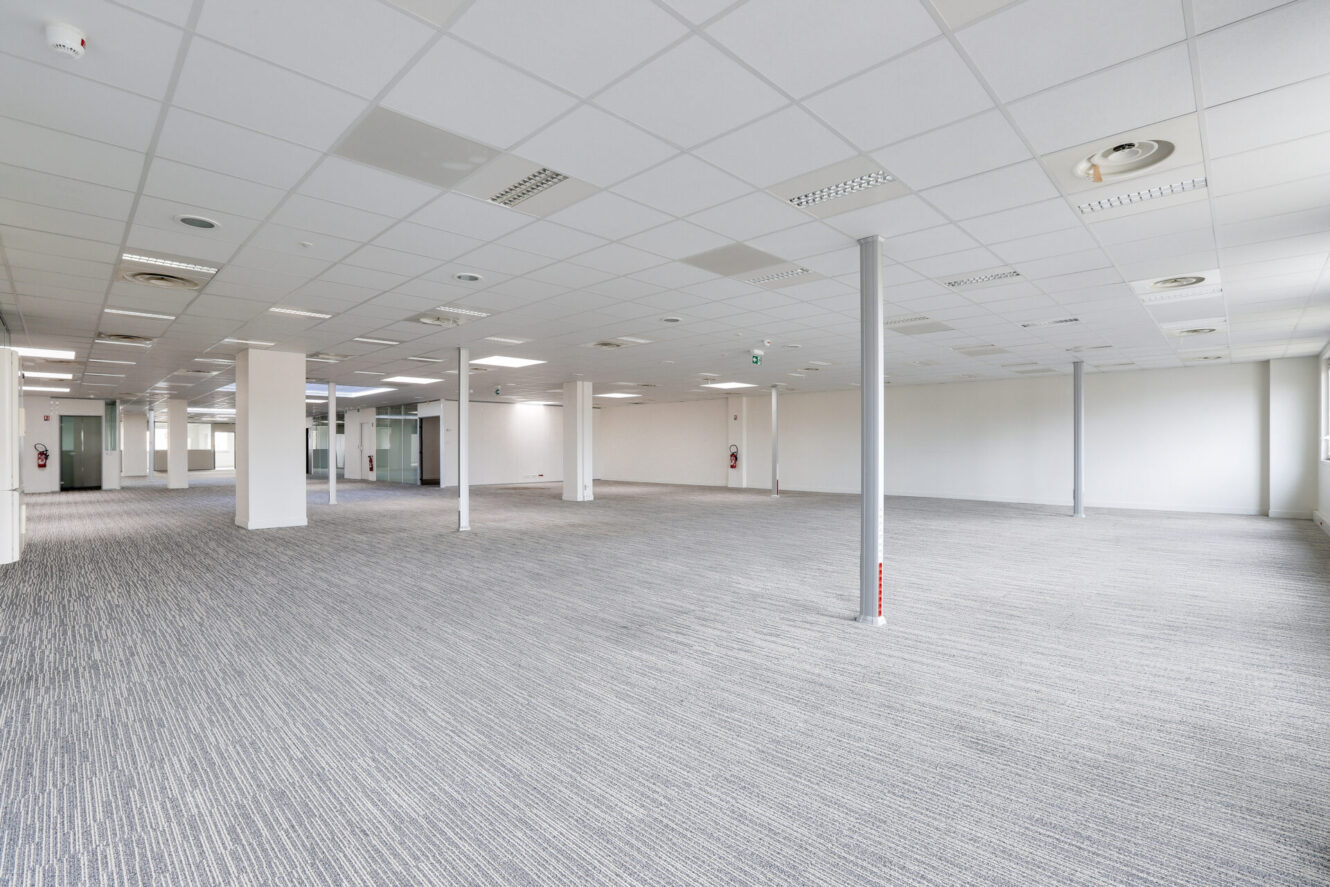 Un bureau spacieux et vide avec des murs blancs, des dalles de plafond, une moquette grise et de multiples colonnes de soutien.