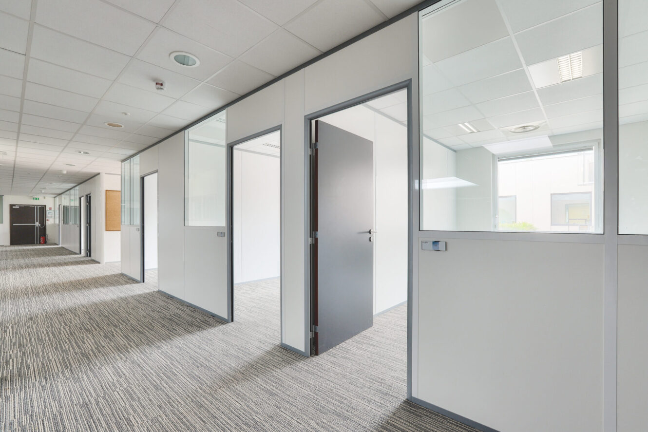 Couloir de bureau moderne vide avec moquette grise, murs blancs, panneaux de verre, portes ouvertes vers les pièces et lumière vive provenant de grandes fenêtres.