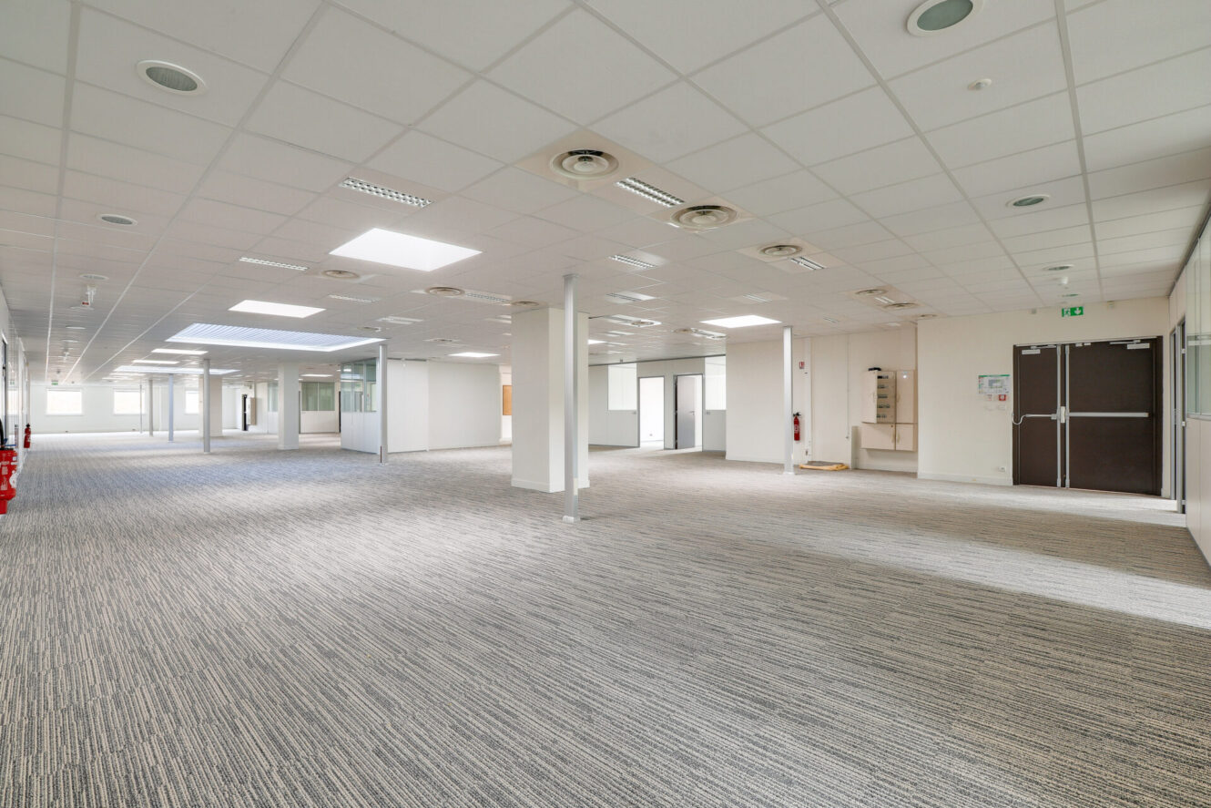 Grand espace de bureau vide avec des sols recouverts de moquette, des murs blancs, des plafonniers et de nombreuses fenêtres ; des doubles portes et une sortie sont visibles.