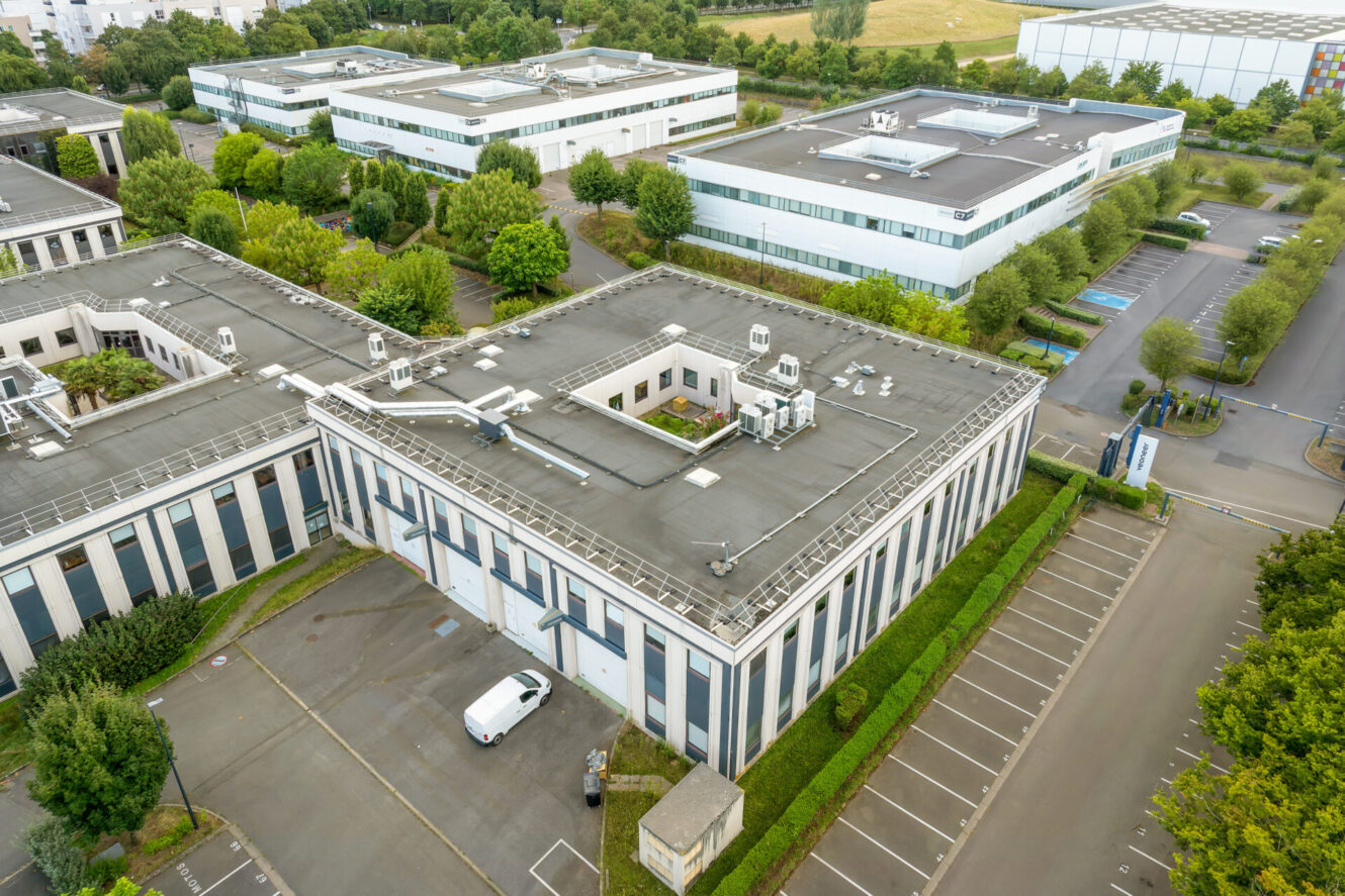 Vue aérienne d'un complexe de bureaux moderne composé de plusieurs bâtiments rectangulaires, de parkings, d'arbres et d'espaces paysagers, par temps nuageux.