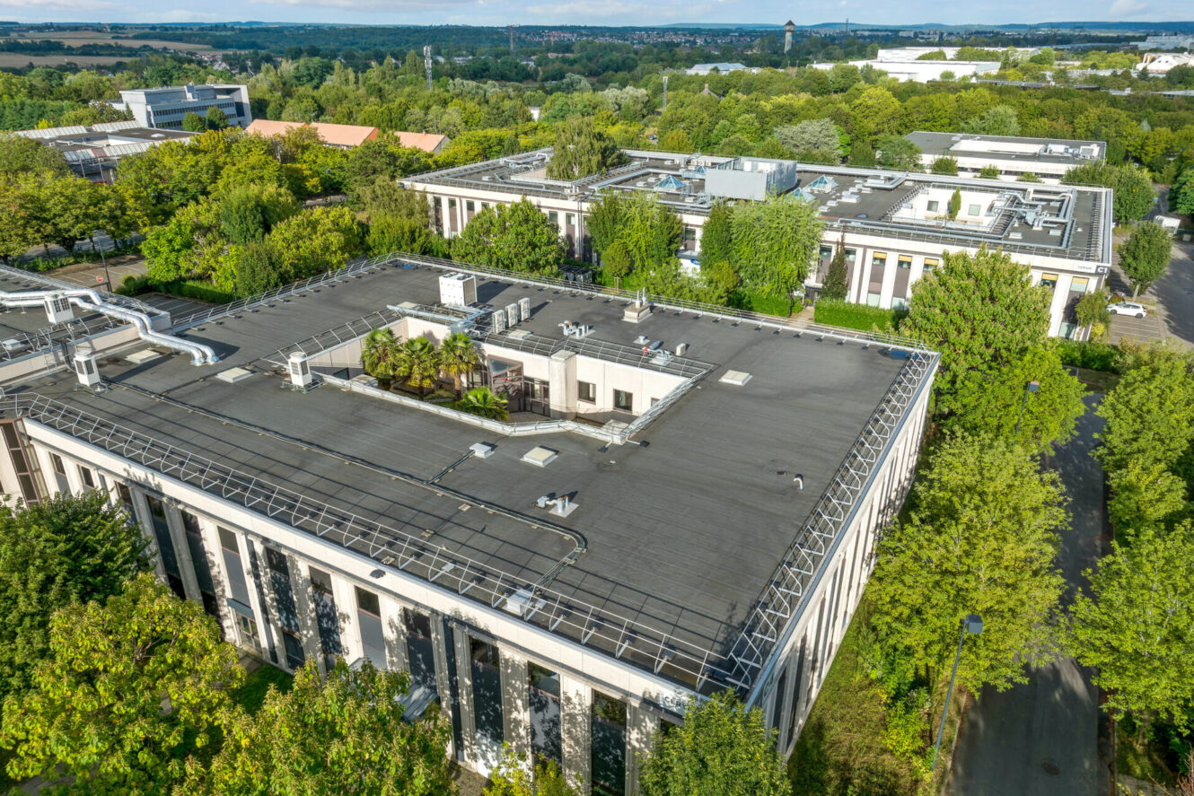 Vue aérienne d'immeubles de bureaux modernes entourés d'arbres, de toits avec des unités de chauffage, de ventilation et de climatisation, et des routes avoisinantes par temps clair.