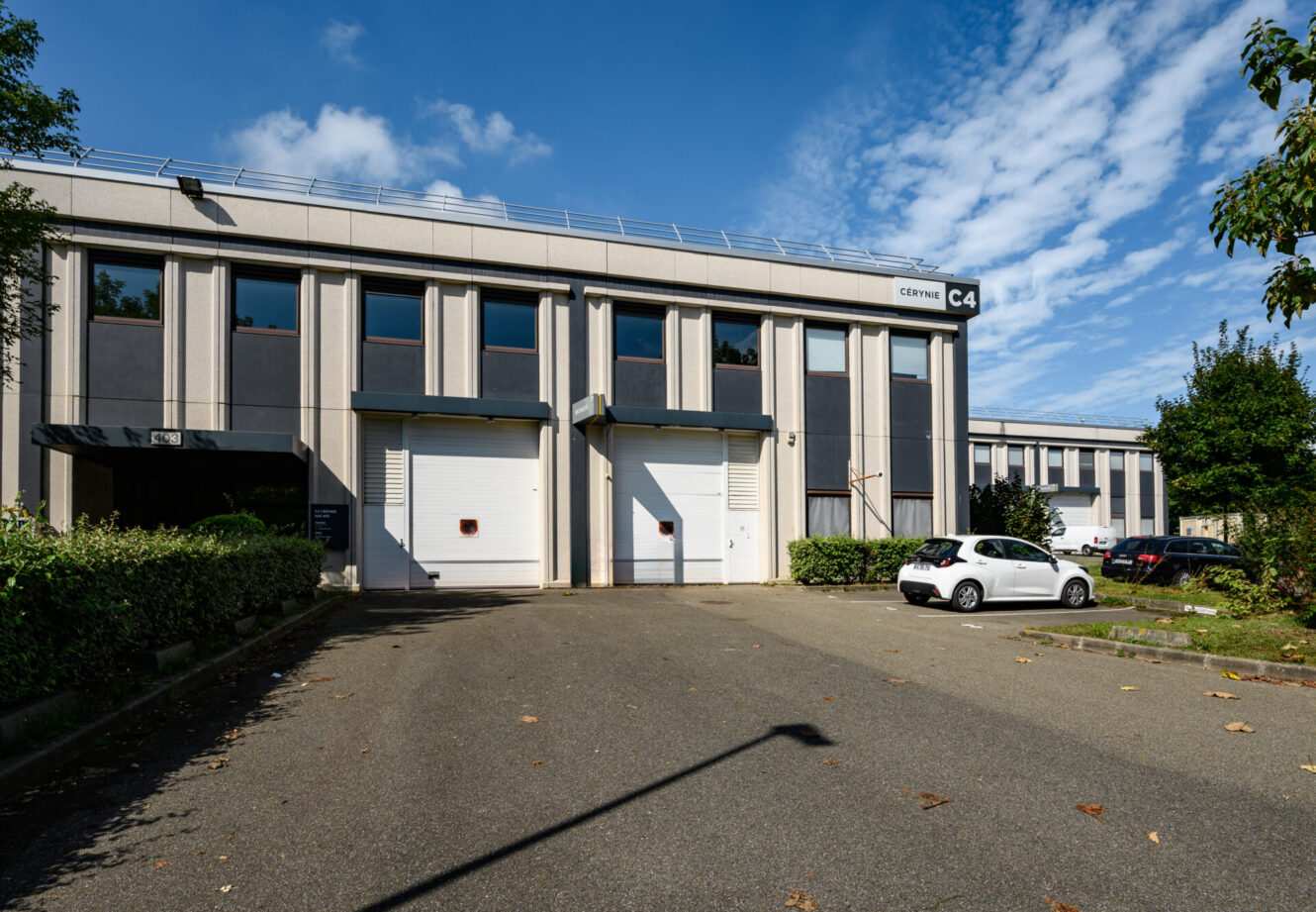 Un bâtiment industriel de deux étages avec de grandes portes de garage blanches portant l'inscription CENTRE C4, des voitures garées à l'extérieur par une journée ensoleillée.