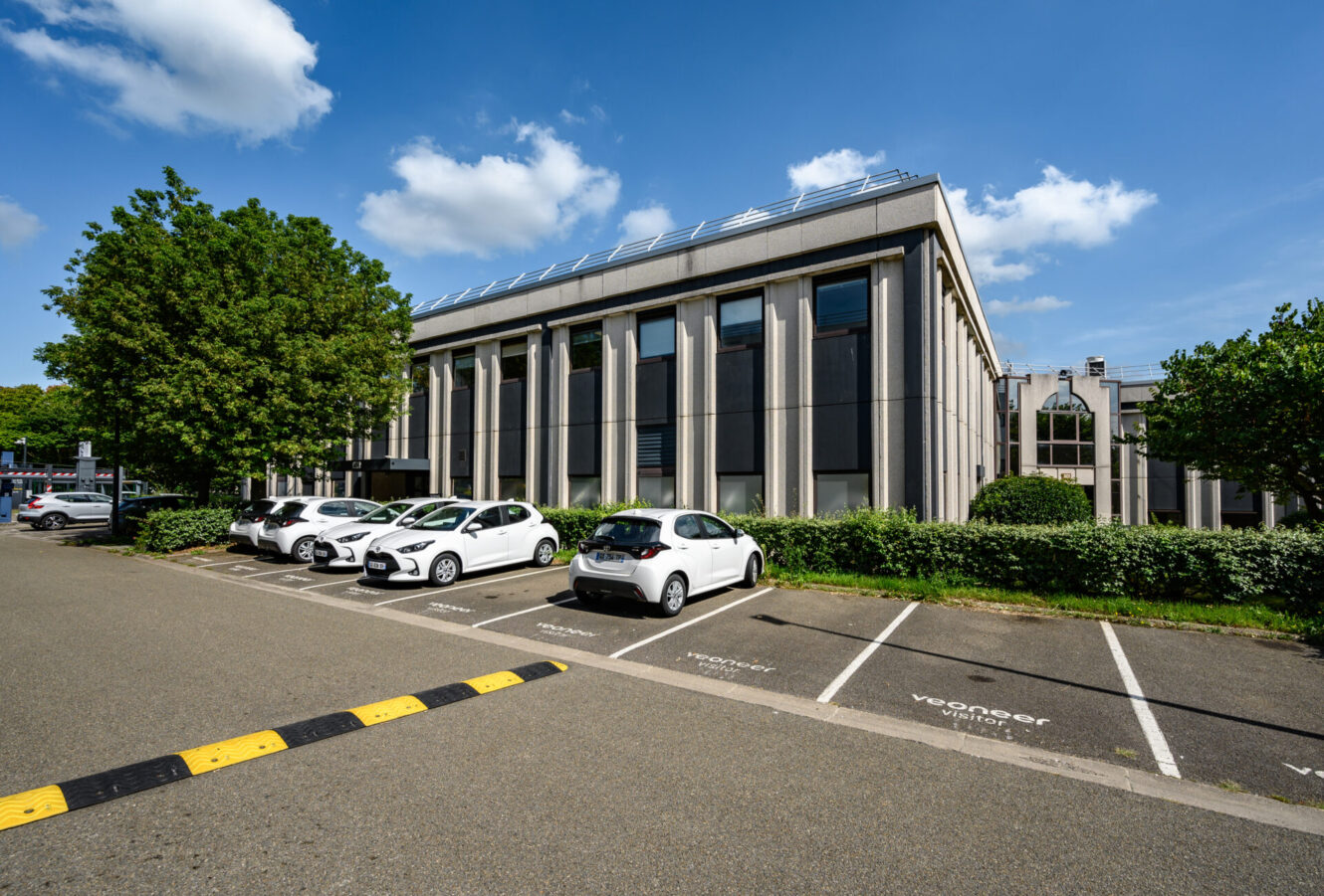 Un immeuble de bureaux moderne avec de grandes fenêtres, des arbres autour et des voitures blanches garées dans le parking voisin sous un ciel bleu.