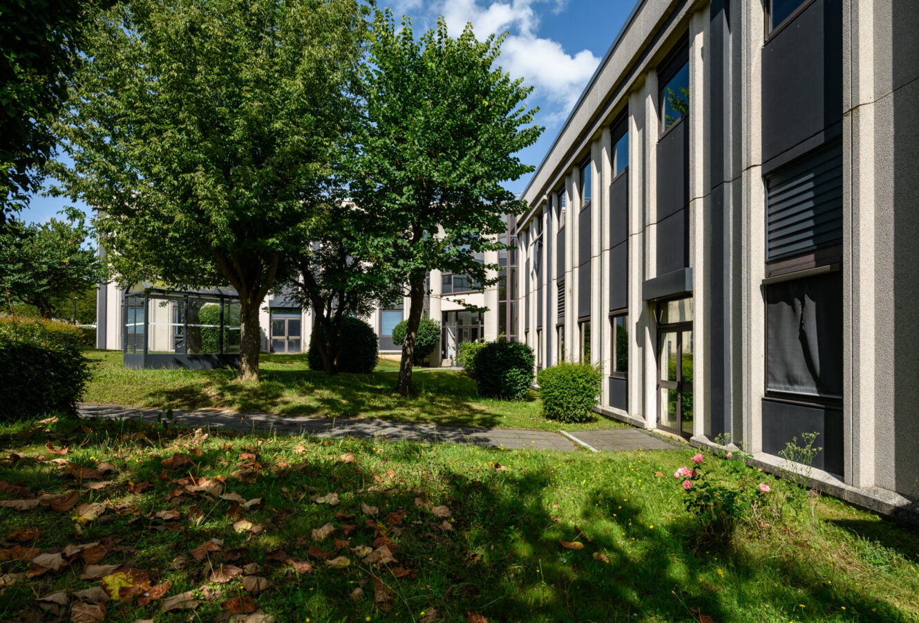 Immeuble de bureaux moderne de deux étages avec de grandes fenêtres, des arbres et des buissons taillés à l'extérieur, sous un ciel bleu avec des nuages épars.