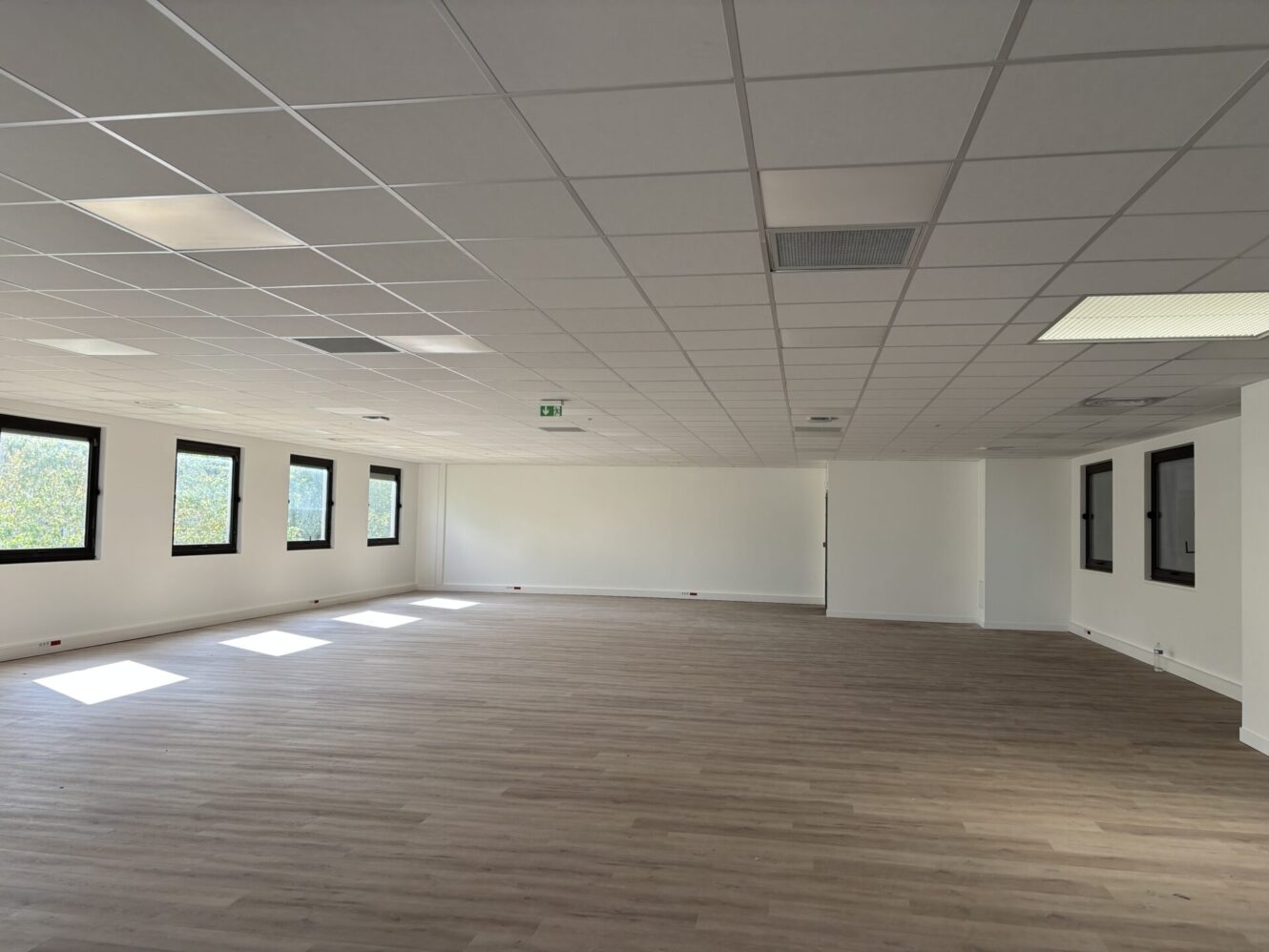 Grand espace de bureau vide avec des murs blancs, des dalles de plafond, plusieurs fenêtres le long d'un mur et un sol en bois clair.