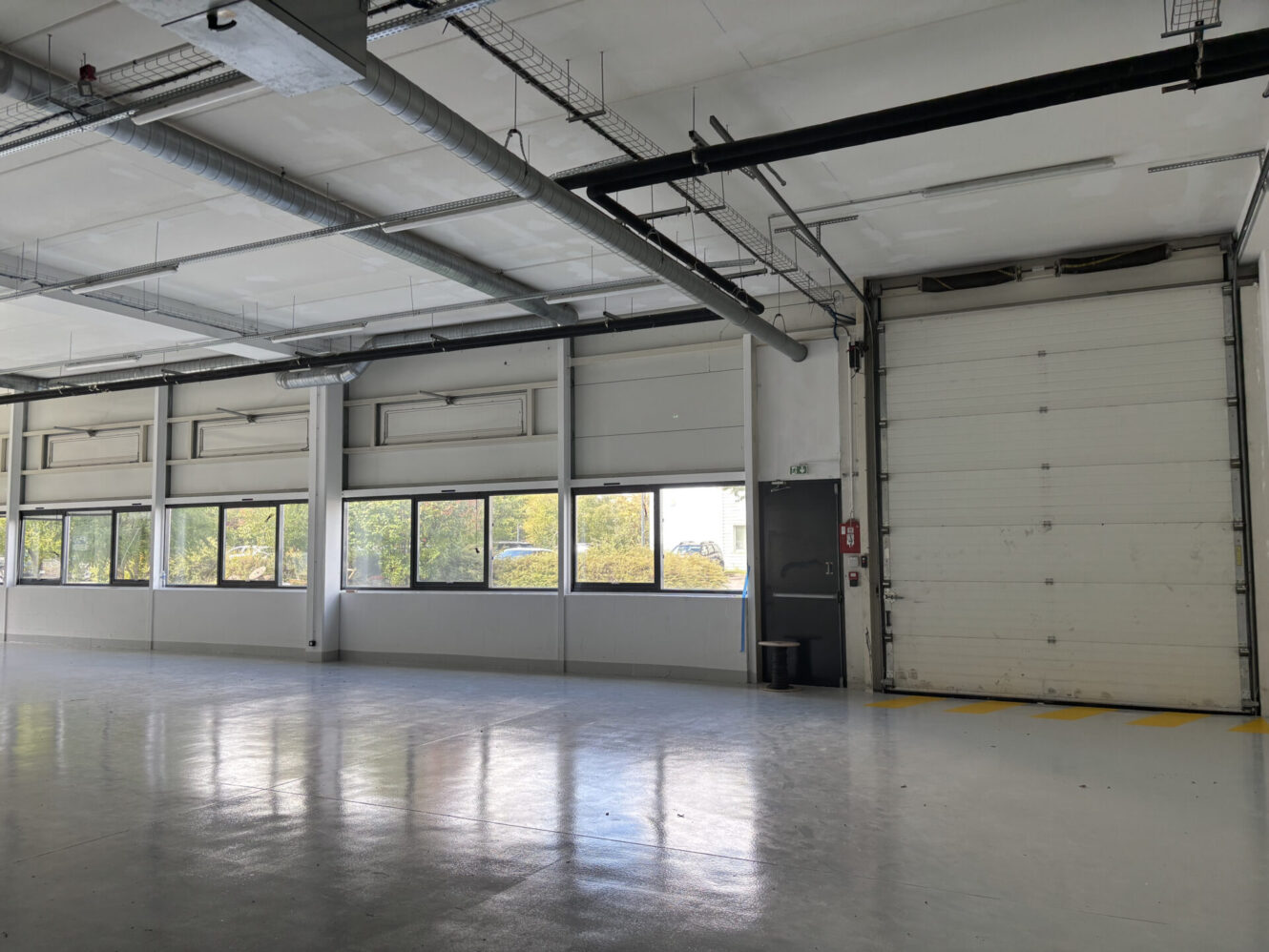 Grand entrepôt industriel vide avec des murs blancs, plusieurs fenêtres, des tuyaux apparents et une porte de garage fermée éclairée par la lumière du jour.
