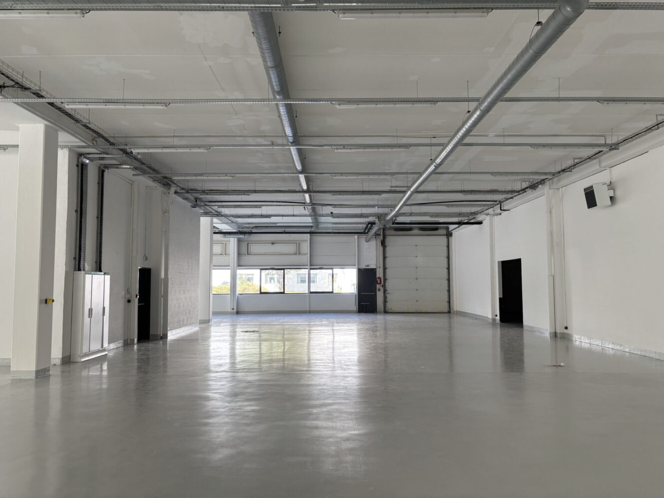 Grand entrepôt industriel vide avec des murs blancs, un sol en béton poli, des tuyaux apparents, des fenêtres et une grande porte roulante.