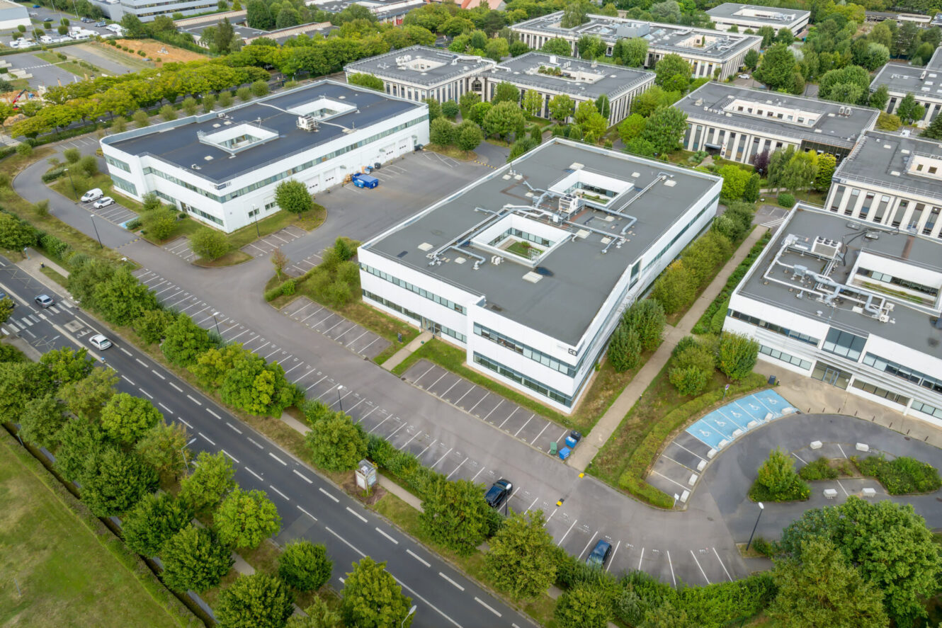 Vue aérienne d'immeubles de bureaux modernes aux toits plats, de parkings, de routes et d'arbres verts entourant la zone.
