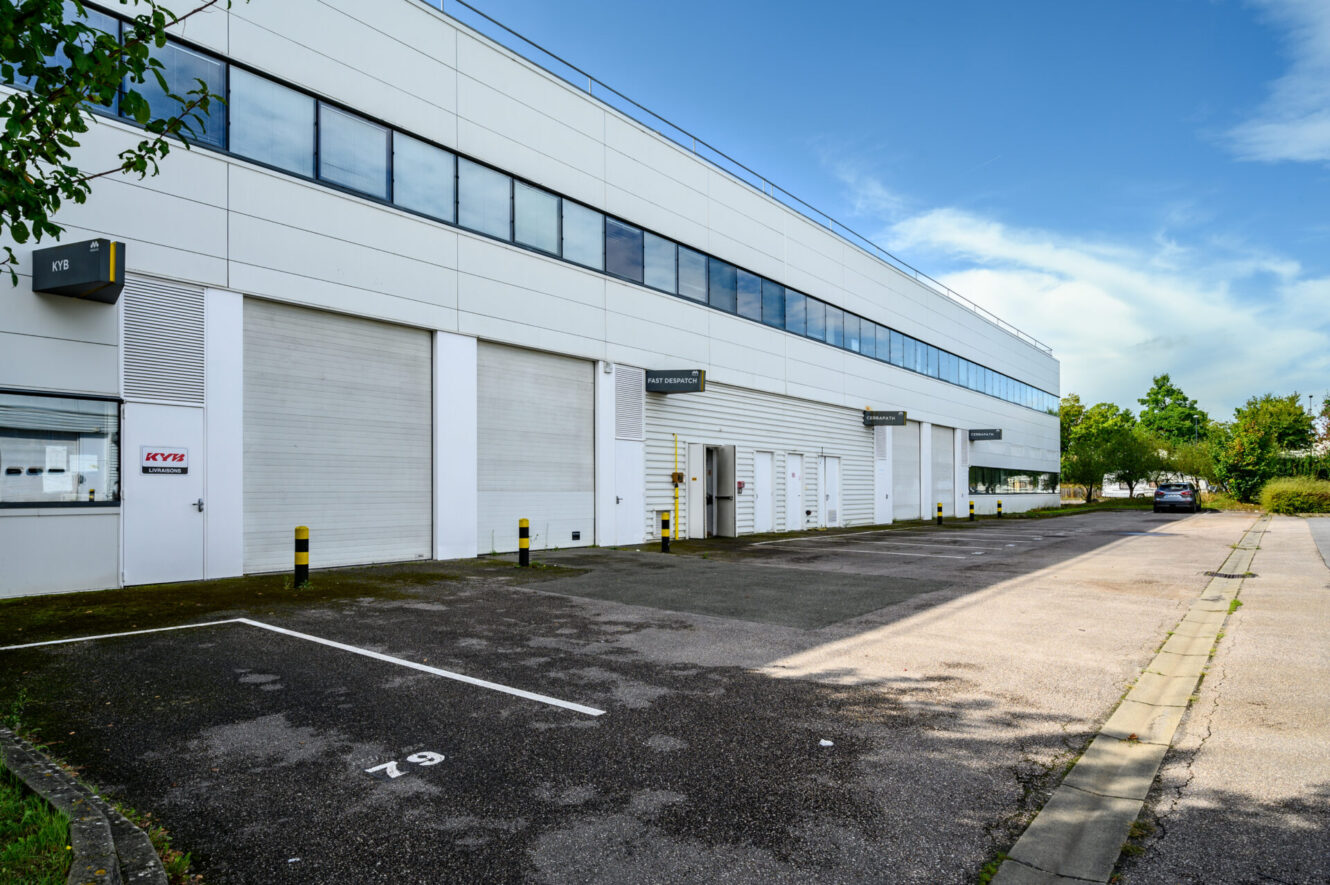 Un bâtiment industriel blanc avec des quais de chargement, des places de parking numérotées, un ciel dégagé et des arbres en bordure de propriété.
