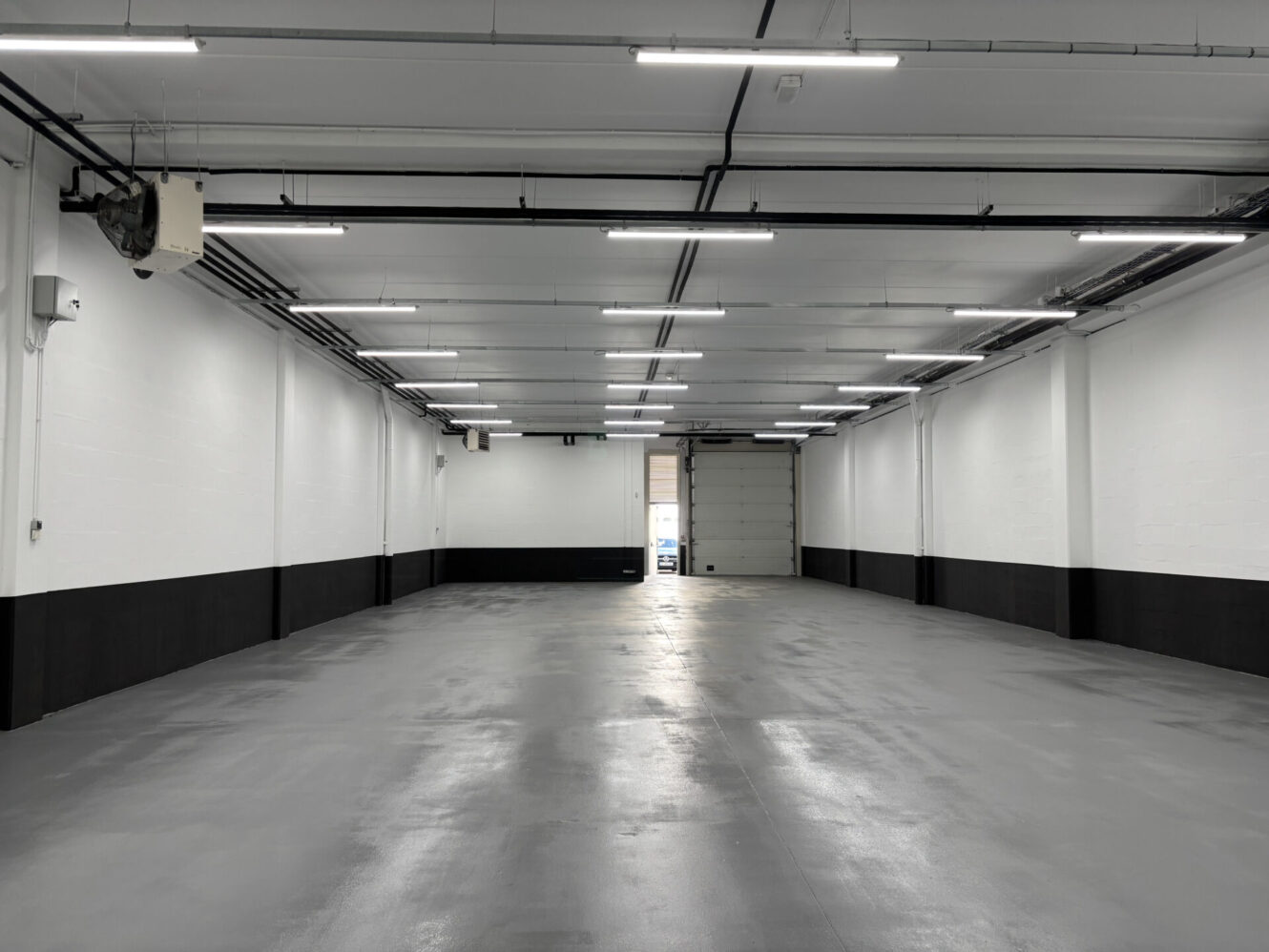 Garage industriel vide et propre avec des murs blancs, des garnitures noires, un éclairage zénithal et une grande porte roulante ouverte à l'extrémité.