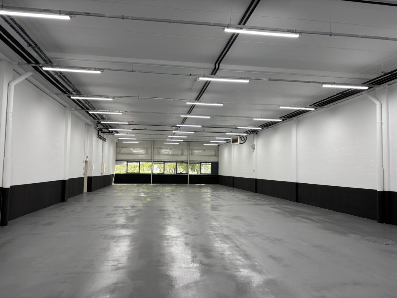 Entrepôt industriel vide et propre, avec un sol en béton poli, des murs blancs, des lampes fluorescentes et de grandes fenêtres laissant entrer la lumière naturelle.
