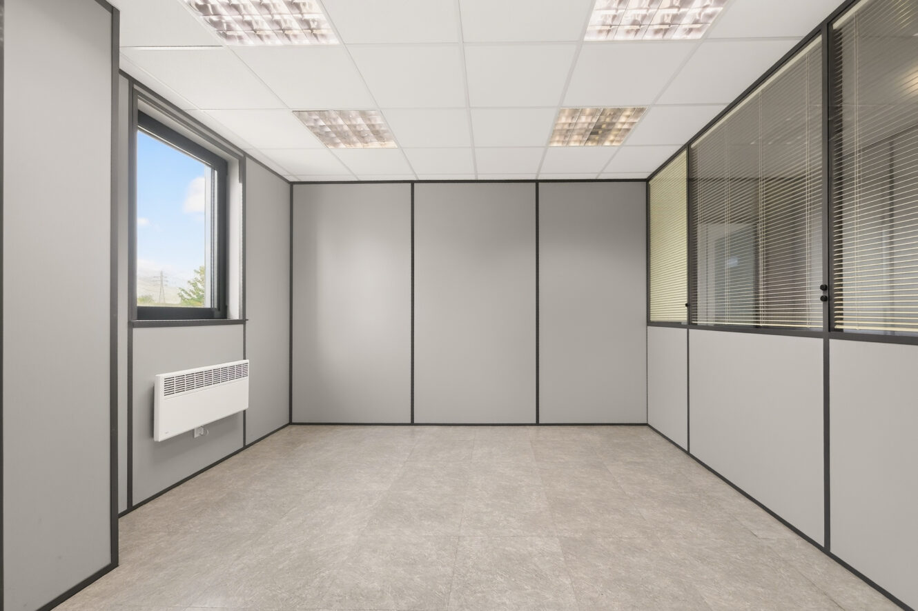 Bureau moderne vide avec des murs gris, un sol carrelé, une fenêtre et un radiateur à gauche, ainsi que des cloisons vitrées équipées de stores à droite.