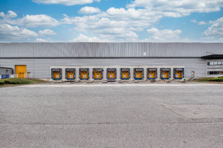 Un grand entrepôt doté de neuf portes de chargement, chacune ornée d'un panneau jaune, et une aire pavée vide devant celui-ci, sous un ciel partiellement nuageux.