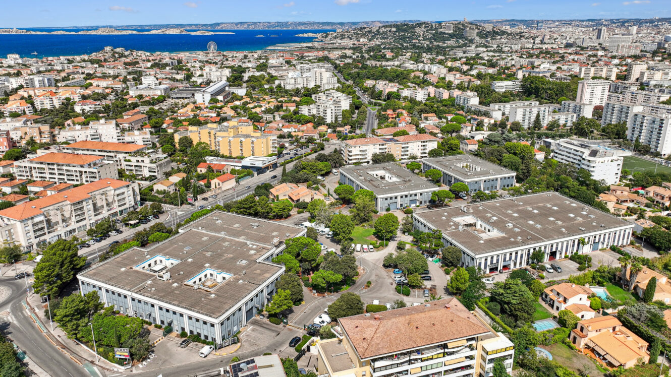 Vue aérienne d'une ville où l'on distingue plusieurs grands immeubles de bureaux rectangulaires, entourés de quartiers résidentiels, d'arbres et d'un littoral lointain, sous un ciel partiellement nuageux.