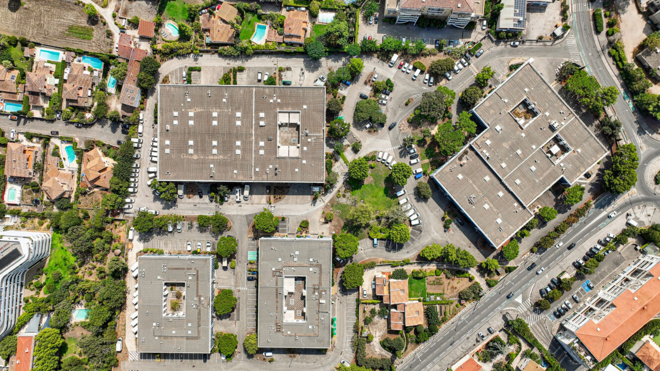 Vue aérienne de trois grands bâtiments rectangulaires dotés de parkings, entourés de maisons avec piscine, d'arbres et de routes qui se croisent.