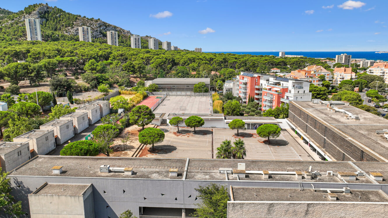 Vue aérienne d'un complexe scolaire ou institutionnel composé de plusieurs bâtiments, de parkings et d'arbres, avec en arrière-plan des collines boisées et, au loin, les gratte-ciel d'une ville située près de la côte.