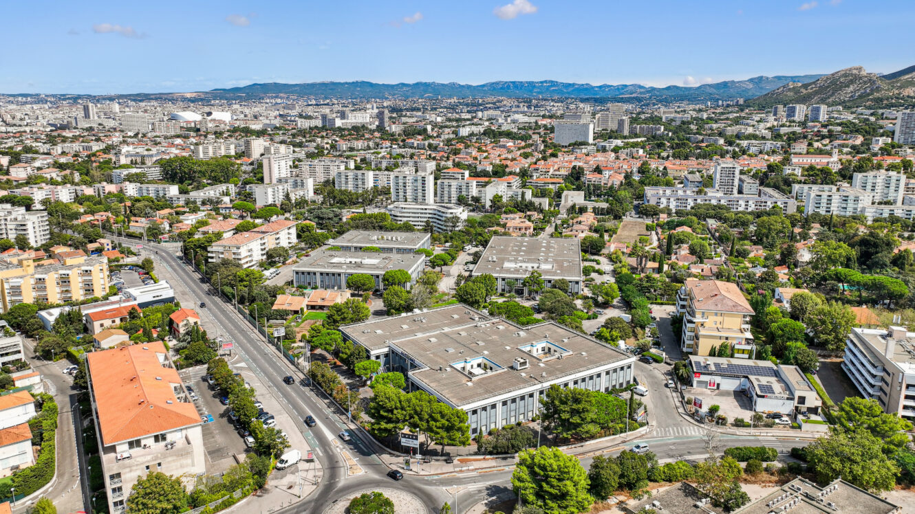 Vue aérienne d'un paysage urbain composé d'immeubles résidentiels, de routes, d'espaces verts et de collines au loin, sous un ciel bleu limpide.