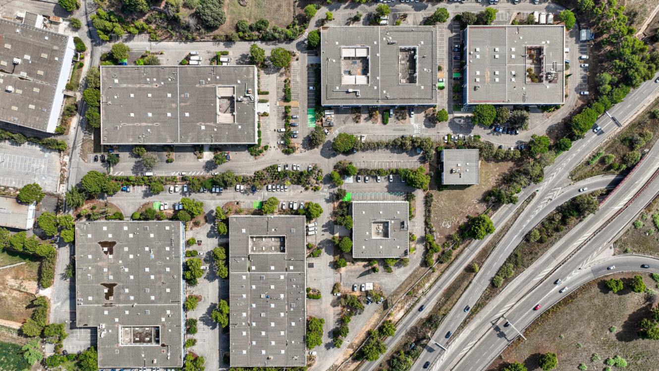 Vue aérienne d'un parc d'activités comprenant des immeubles de bureaux, des parkings et un espace vert luxuriant, situé à proximité d'une bretelle d'autoroute.