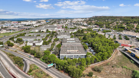 Vue aérienne d'une zone industrielle et commerciale comprenant plusieurs bâtiments, des routes environnantes, une végétation dense et un paysage urbain au loin, sous un ciel bleu parsemé de nuages.