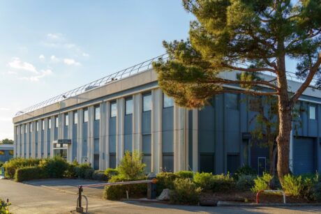 Un immeuble de bureaux moderne à deux étages, doté de hautes fenêtres, est entouré d'arbres et d'arbustes, baigné par la lumière du jour.