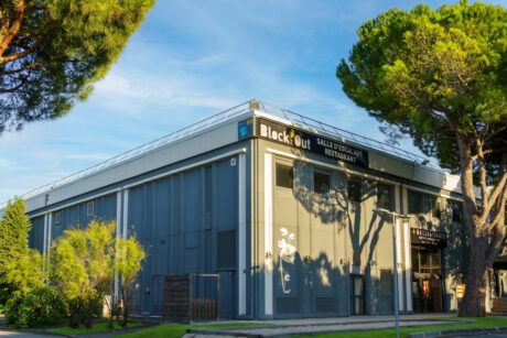 Un bâtiment moderne gris, doté de grandes baies vitrées et arborant une enseigne « BlockOut », entouré d'arbres et de verdure sous un ciel dégagé.