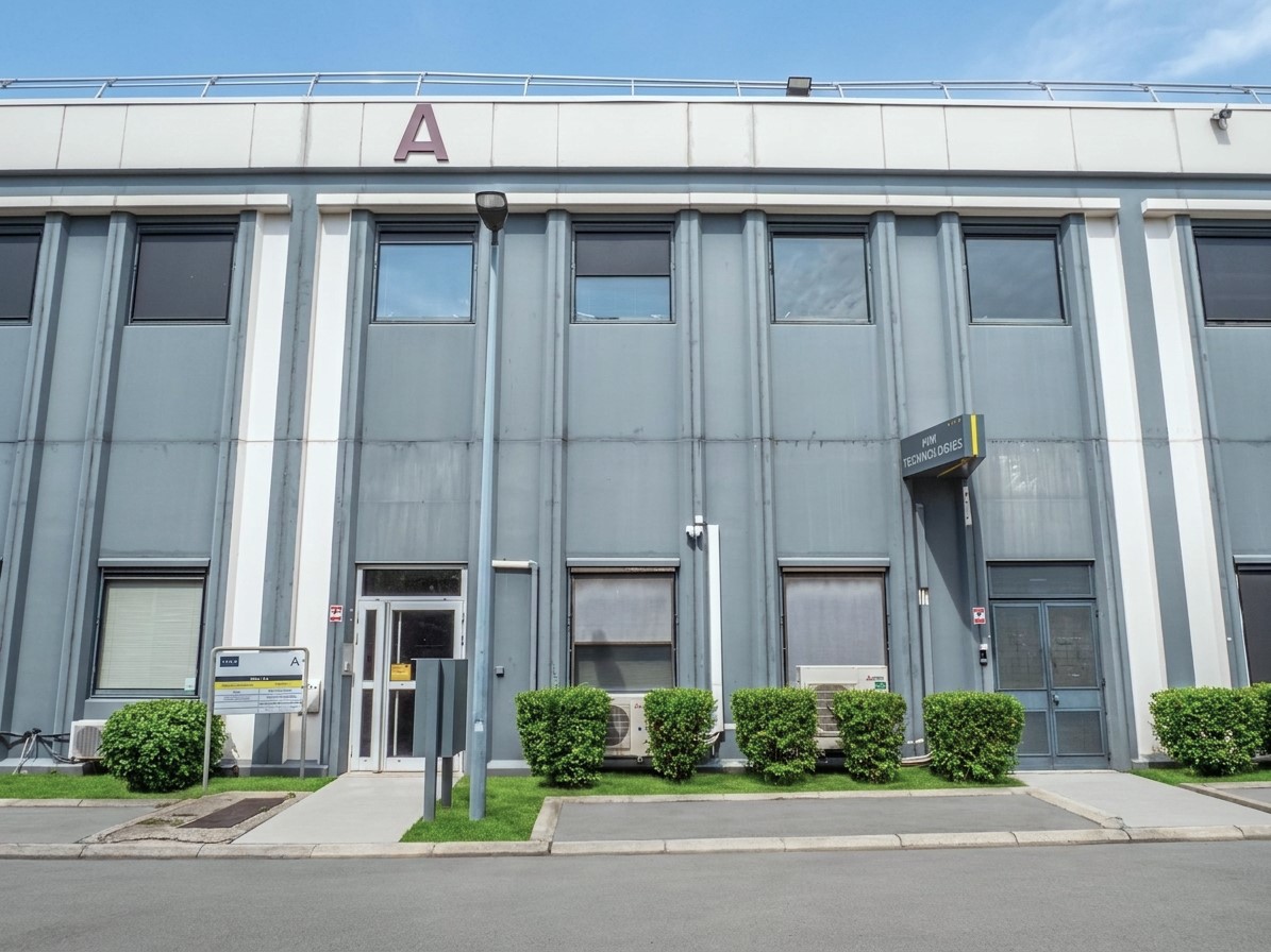 Un bâtiment industriel gris, doté de hautes fenêtres, d'une porte et d'une enseigne portant l'inscription « A. ». Des buissons bordent le trottoir devant le bâtiment, sous un ciel bleu limpide.